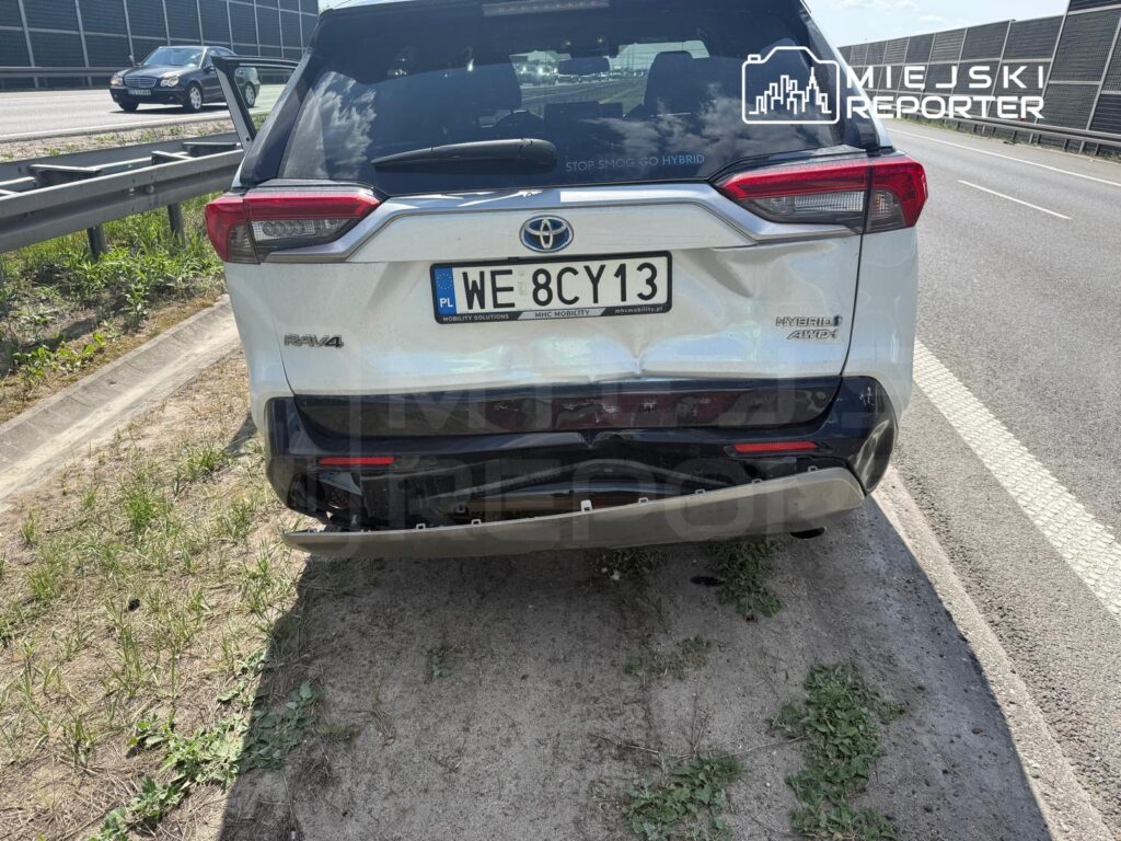Biały samochód osobowy marki Toyota RAV4 zaparkowany na poboczu drogi, z otwartymi drzwiami tylnymi.