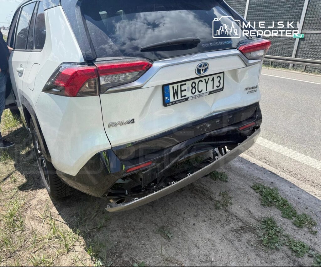 Uszkodzony tył białego samochodu marki Toyota RAV4 stojącego na poboczu drogi, obok mężczyzna w niebieskiej koszuli.