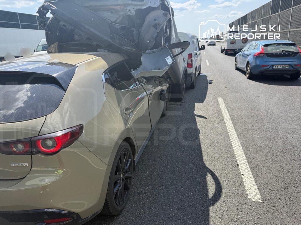 Zniszczony samochód leży na dachu innego pojazdu na ruchliwej drodze, obok stoją inne auta.