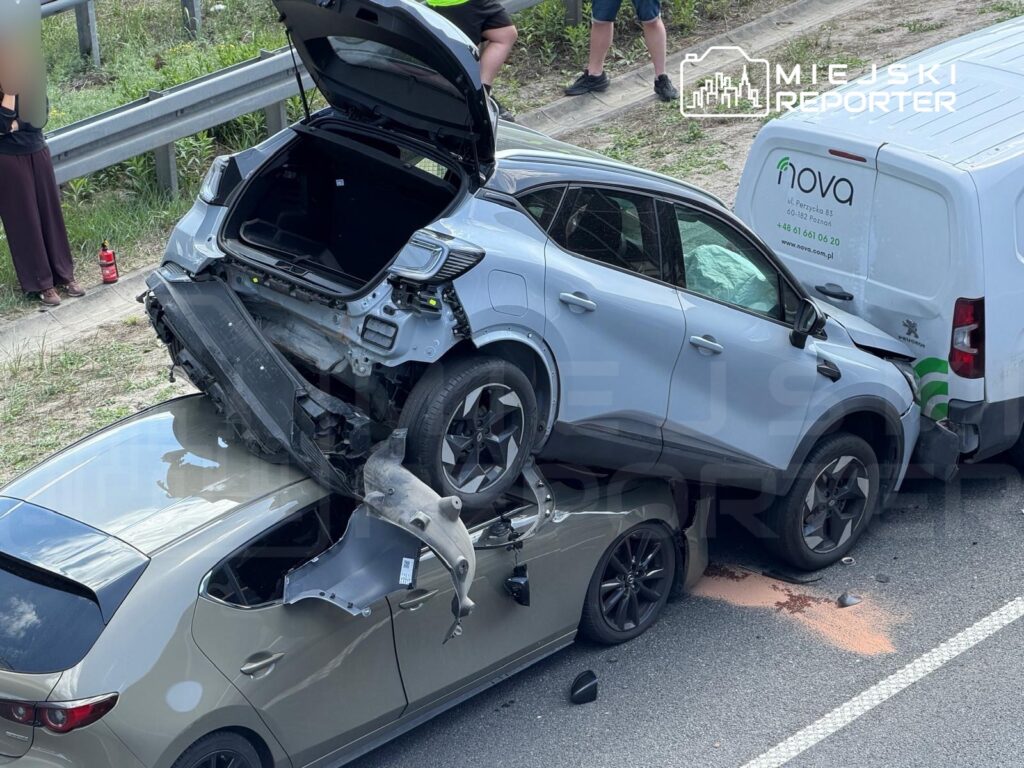 Trzy zderzone samochody na drodze, w tym jeden z uniesioną maską, obok stojący gapie. Na jezdni plama płynów.