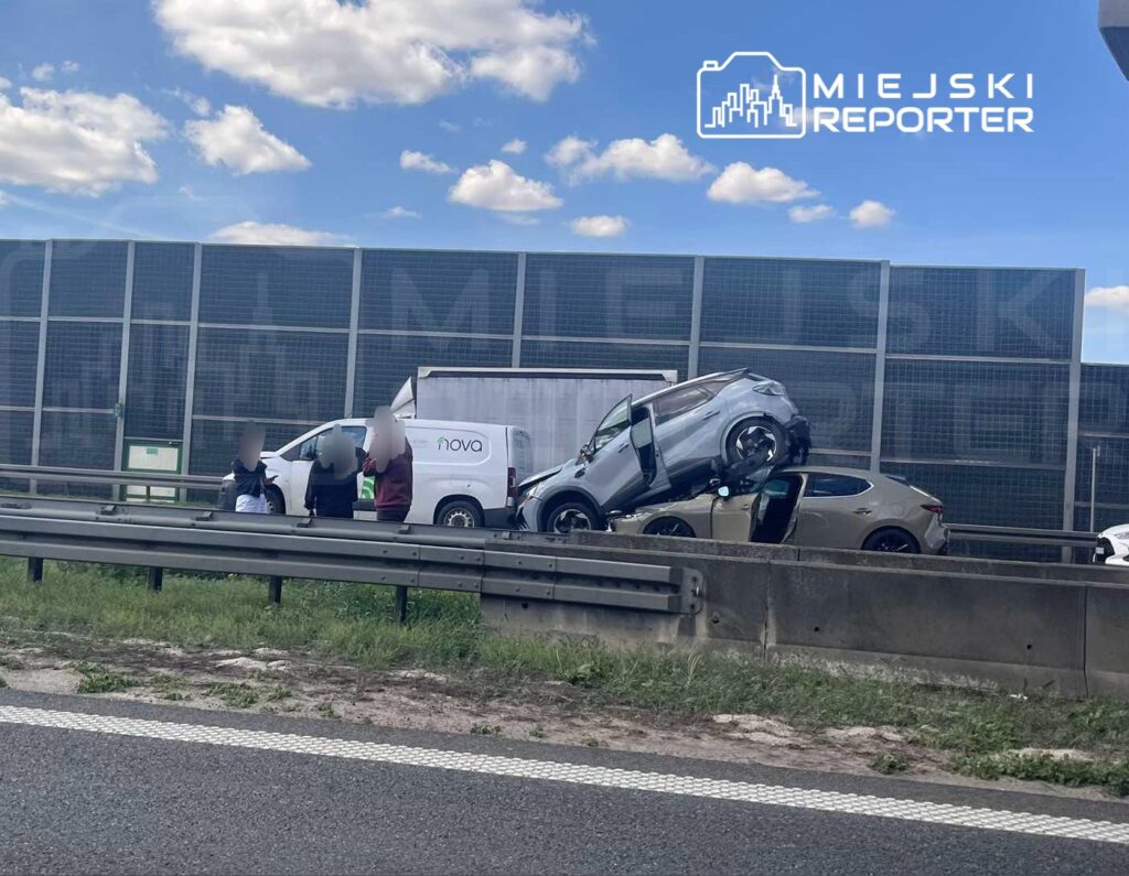 Na drodze stoją dwie uszkodzone samochody, z jednym z nich uniesionym na dachu drugiego, obok grupka osób.