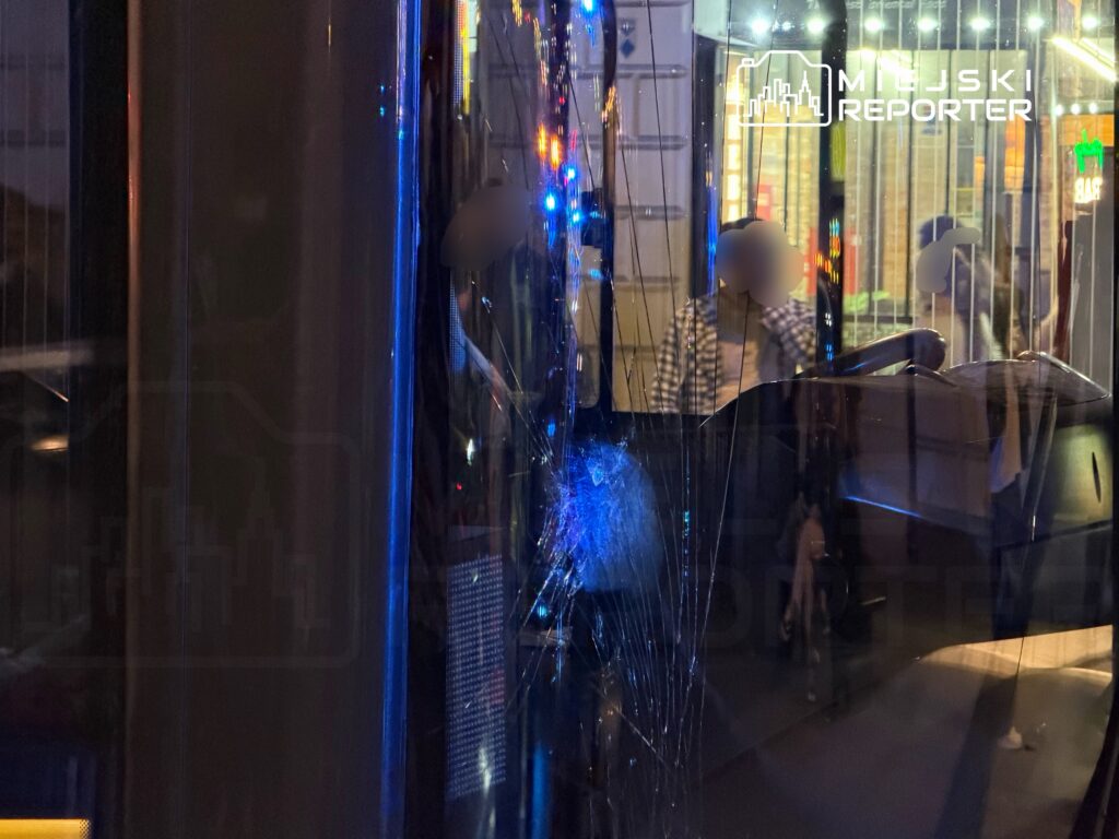 Pęknięta szyba w drzwiach pojazdu, w tle widać ludzi i oświetlone budynki nocą.