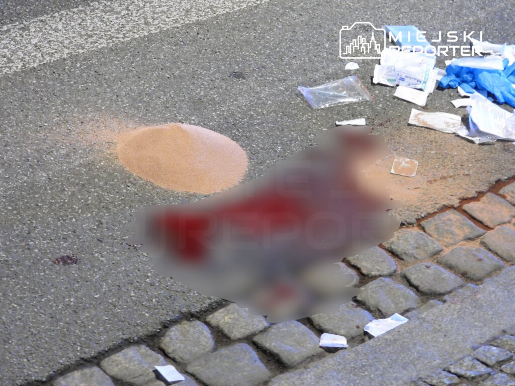 Na jezdni widać kopczyk piasku oraz rozrzucone opakowania medyczne obok plamy.