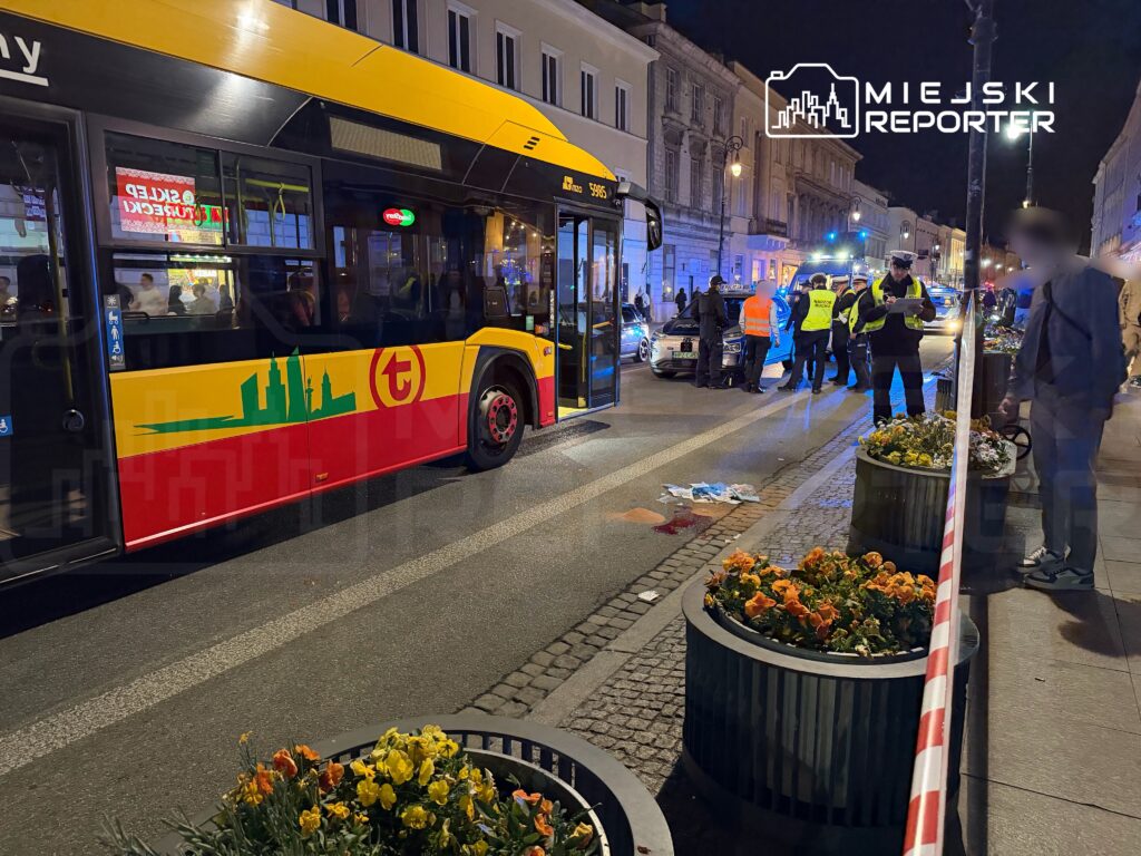Na ulicy widoczny jest autobus komunikacji miejskiej, a w tle policjanci prowadzący dochodzenie w miejscu zdarzenia.