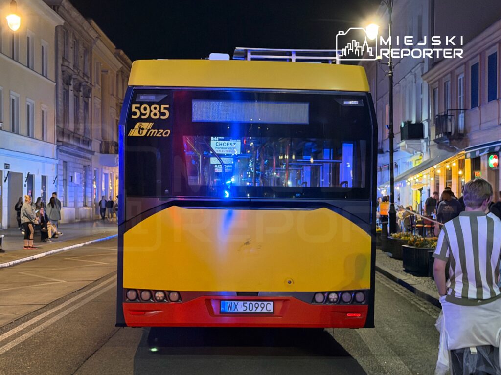 Jasnożółty autobus z numerem taborowym 5985 stoi na miejskiej ulicy w nocy, otoczony przez pieszych i oświetlenie uliczne.
