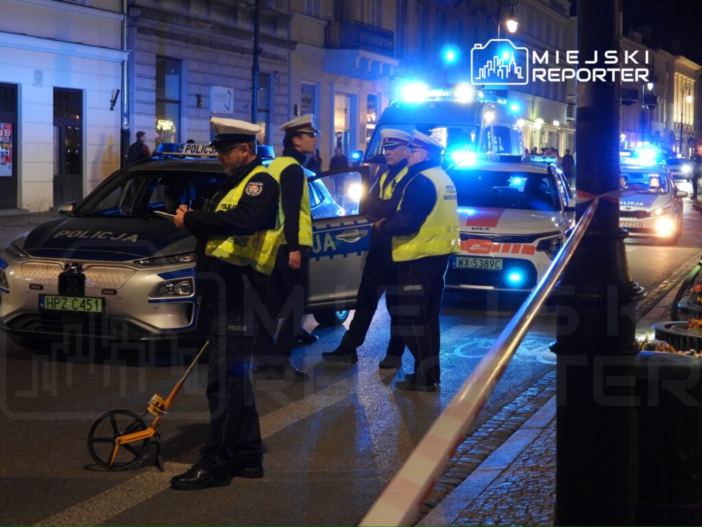 Policjanci w kamizelkach odblaskowych prowadzą czynności dochodzeniowe na oświetlonej ulicy, obok radiowozów.