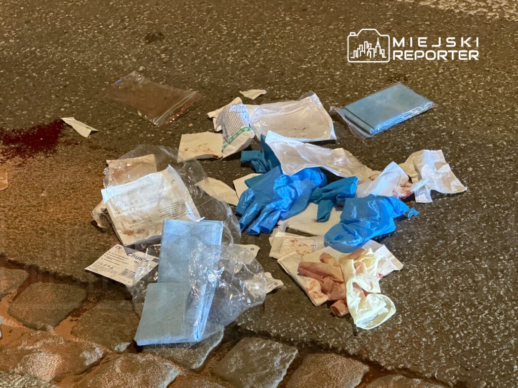 Na nawierzchni drogi leżą porozrzucane opakowania, rękawiczki i papierowe materiały, z plamami krwi w tle.