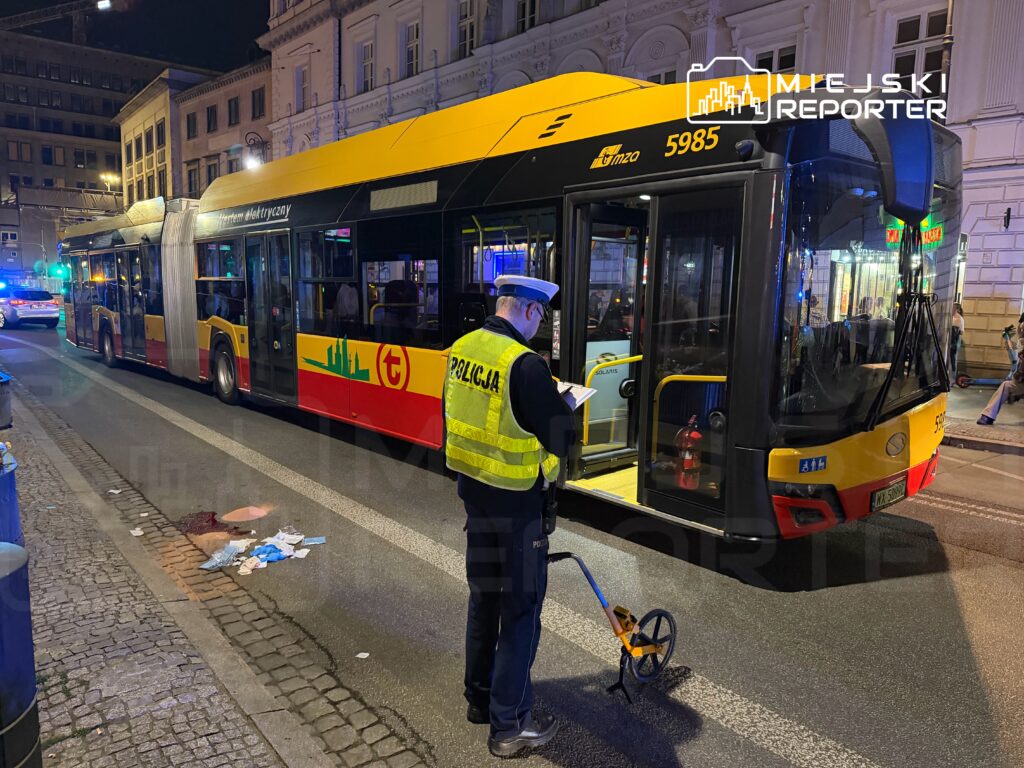 Policjant w kamizelce odblaskowej mierzy odległość przy elektrycznym autobusie na oświetlonej ulicy, obok leżące ubrania.