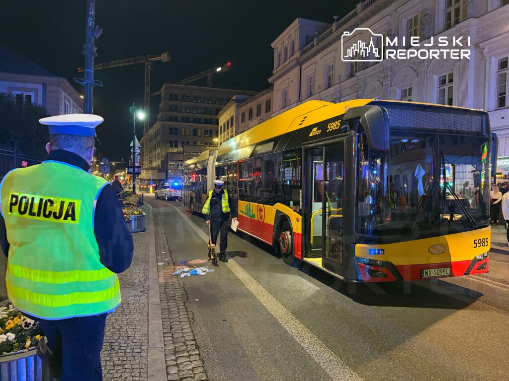 Policjant w neonowej kamizelce obserwuje interwencję przy przystanku autobusowym nocą, obok stoi autobus komunikacji miejskiej.