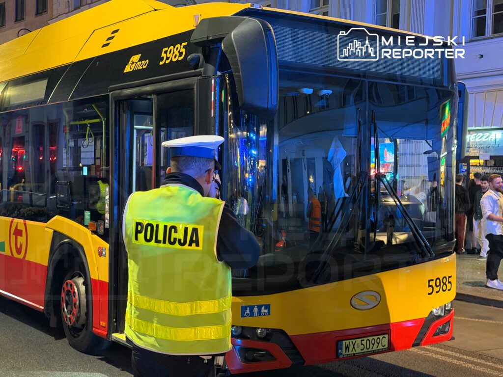 Policjant w odblaskowej kamizelce obserwuje zatrzymany autobus na ruchliwej ulicy nocą.