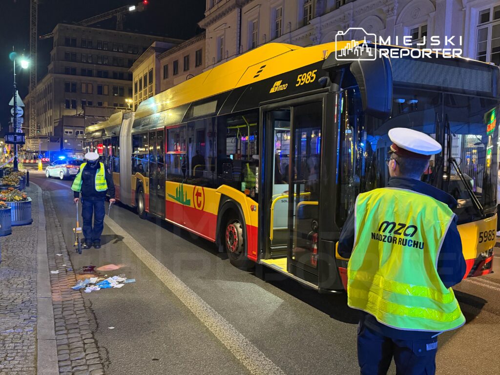 Funkcjonariusz w kamizelce odblaskowej bada miejsce zdarzenia obok autobusu miejskiego na oświetlonej ulicy.