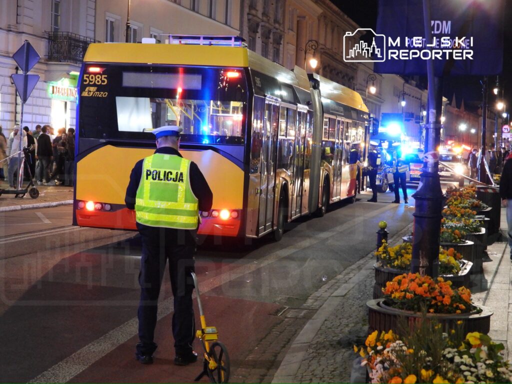Policjant w odblaskowej kamizelce mierzy odległość przy zatrzymanym autobusie w oświetlonej ulicy nocą.
