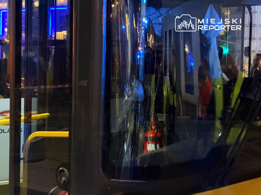 Zniszczona szyba w autobusie, odbijająca światła miejskich neonów i pasażerów w tle.