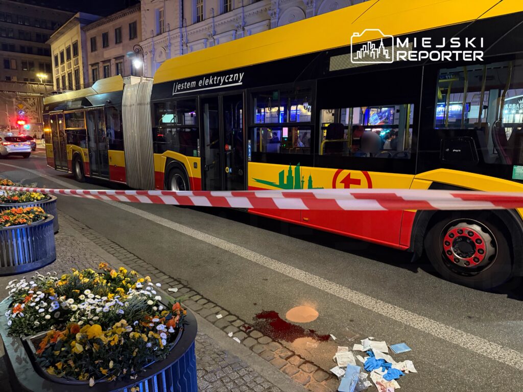 Autobus elektryczny zatrzymany na ulicy, w pobliżu kwietników z kwiatami, obok plama na jezdni i porozrzucane przedmioty.