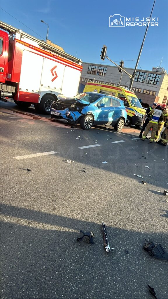 W wyniku wypadku drogowego zderzyły się niebieski samochód osobowy i czerwona ciężarówka strażacka na ulicy, obok której stoją ratownicy medyczni.