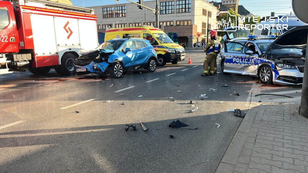 W wyniku wypadku drogowego dwa uszkodzone samochody stoją na jezdni, wokół rozrzucone są fragmenty pojazdów. Obok działa strażak oraz policjant.