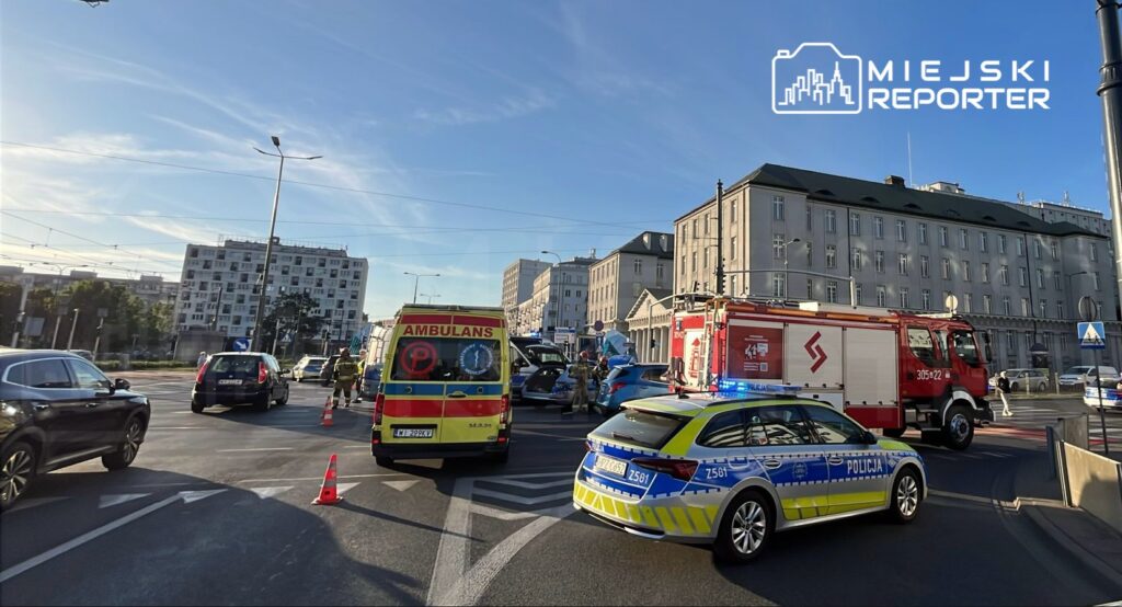 Karetka pogotowia oraz samochód straży pożarnej blokują ruch na ulicy, obok stoją policjanci oraz zablokowane pojazdy.