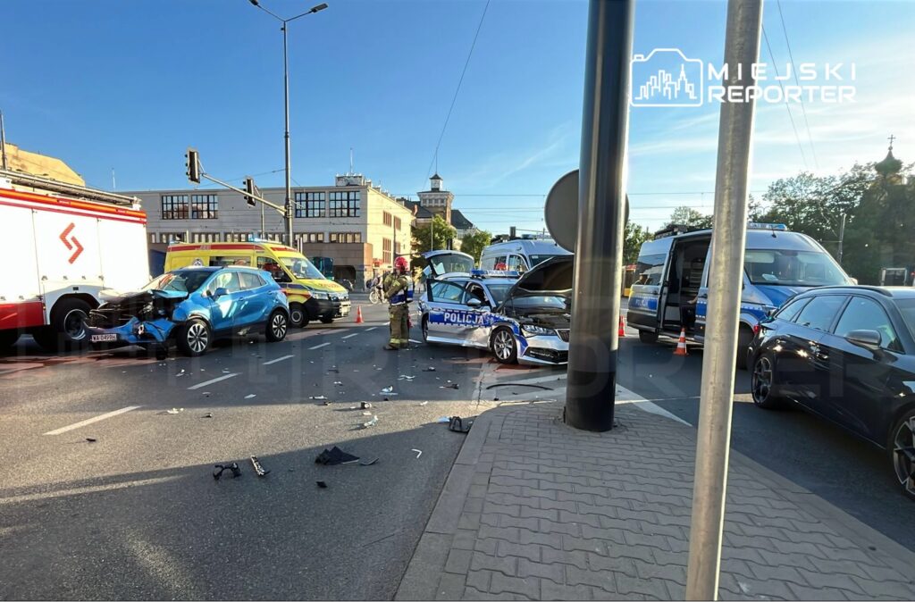 Na skrzyżowaniu znajduje się uszkodzony niebieski samochód osobowy oraz policyjny radiowóz z otwartą maską; w tle widać pojazdy ratunkowe.
