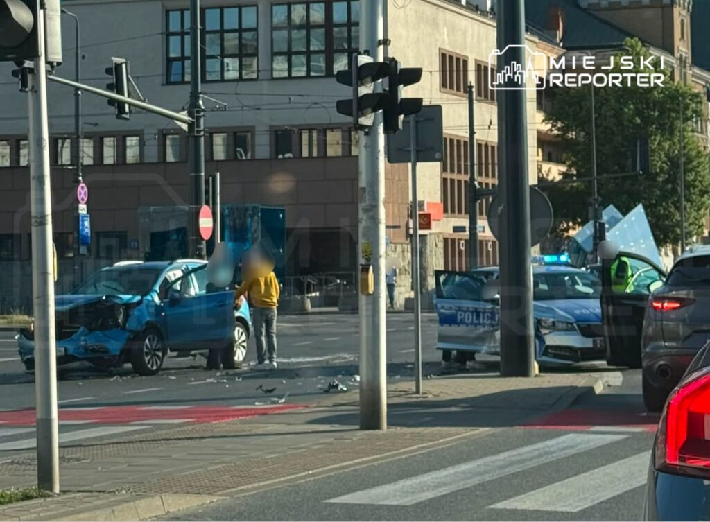 Mężczyzna w żółtej kurtce rozmawia z policjantem obok uszkodzonego niebieskiego samochodu na skrzyżowaniu.