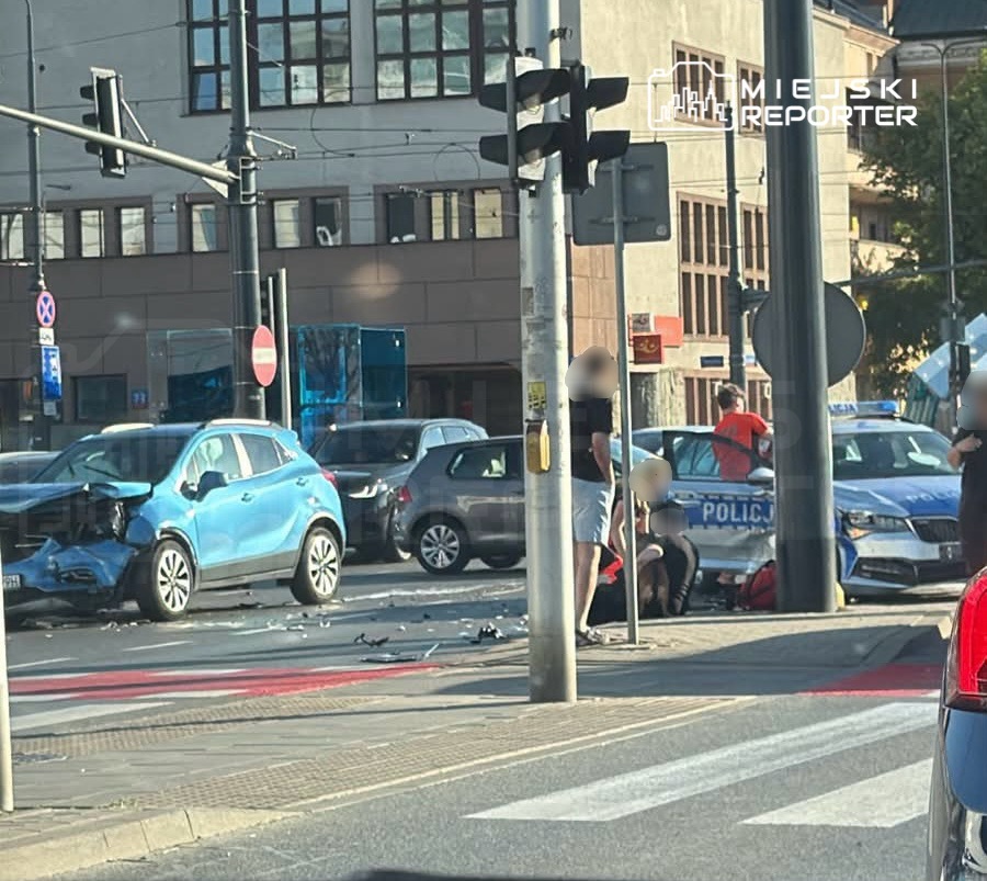 W wyniku kolizji drogowej uszkodzony niebieski samochód stoi na skrzyżowaniu, obok interweniujący policjanci oraz osoby poszkodowane.
