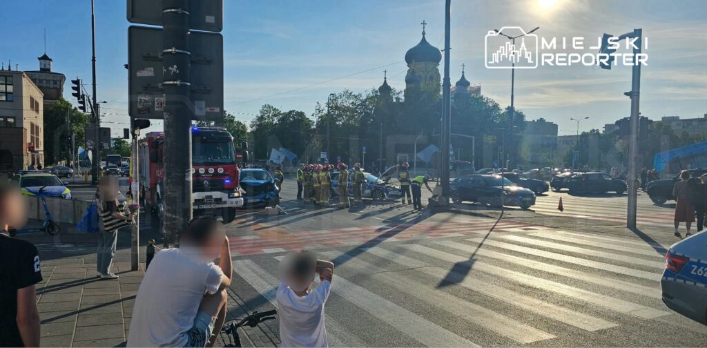 Na skrzyżowaniu ulicy z widocznymi pojazdami, strażacy oraz policja uczestniczą w akcji ratunkowej po wypadku drogowym.
