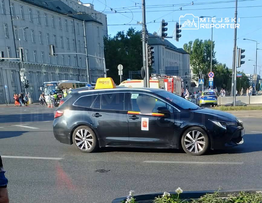 Czarny taksówka skręcająca w ruchliwym skrzyżowaniu, w tle budynki i przechodnie.