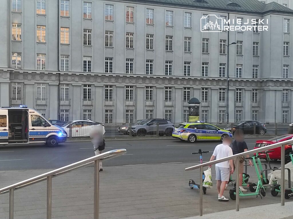 Policyjny radiowóz i karetka stoją na ulicy obok grupy osób z hulajnogami, w tle nowoczesny budynek.