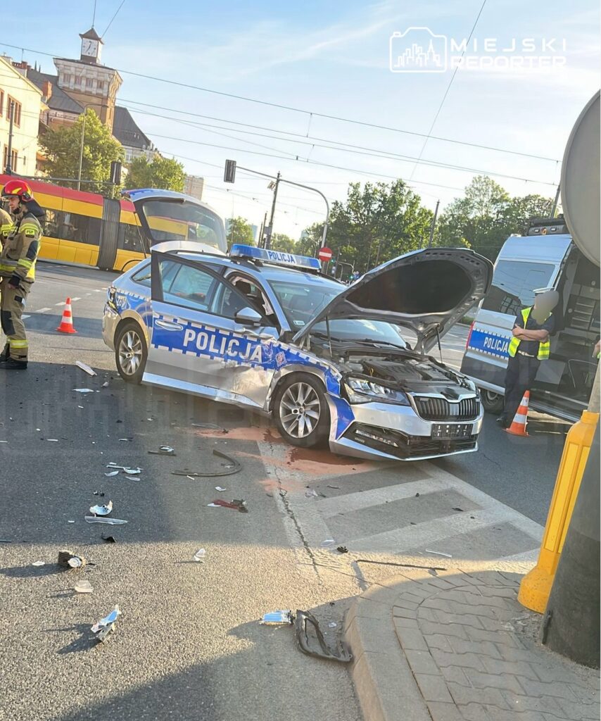 Policyjny samochód z otwartą maską, uszkodzony w wyniku kolizji, stoi na skrzyżowaniu, obok pracownicy służb ratunkowych.
