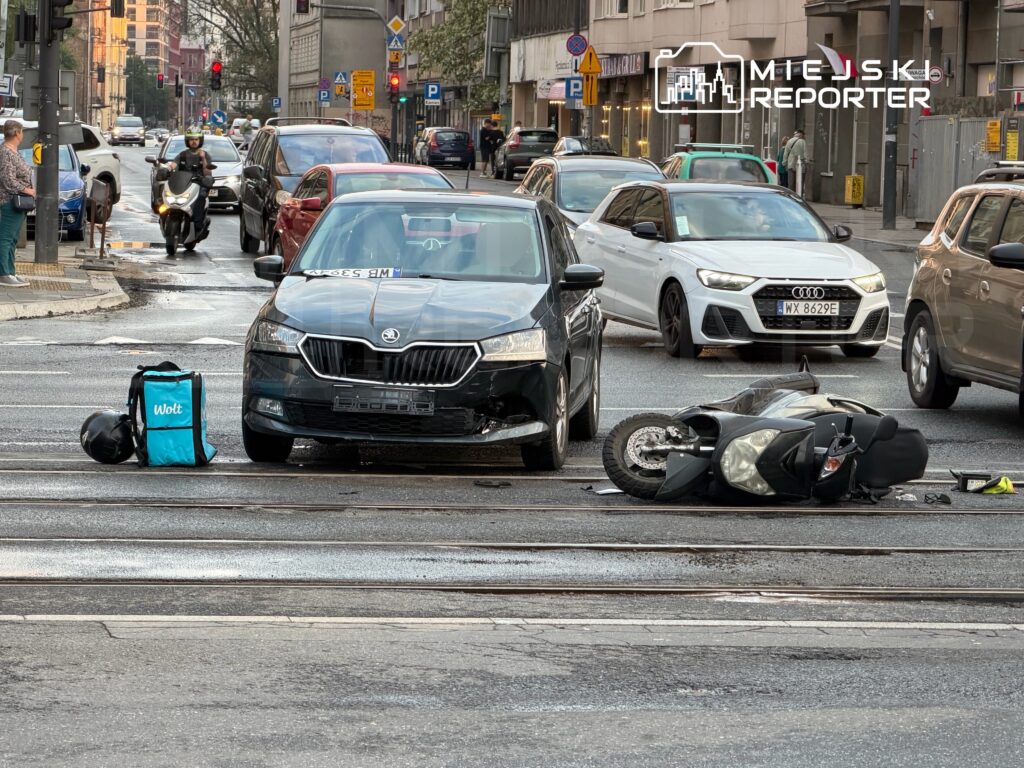 Czarny samochód osobowy stoi na jezdni obok przewróconego motocykla, a obok niego leży torba z logiem Wolt.