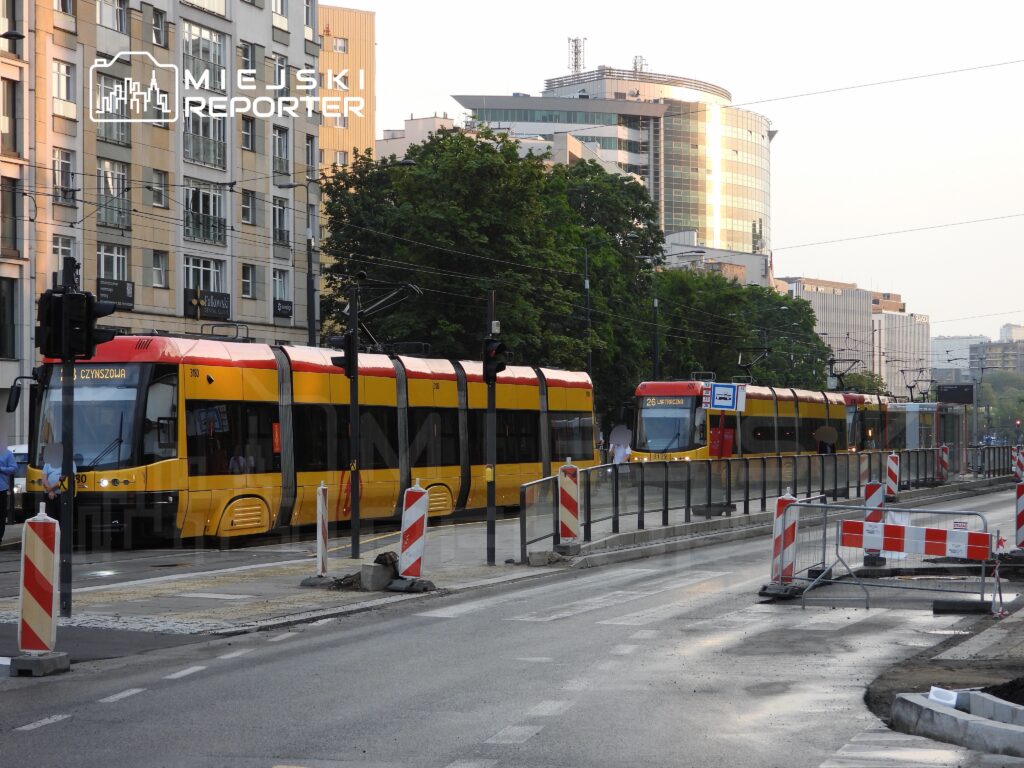 Tramwaje w żółto-czerwonych barwach zatrzymane na przystanku w mieście, w tle nowoczesne budynki i prace drogowe.