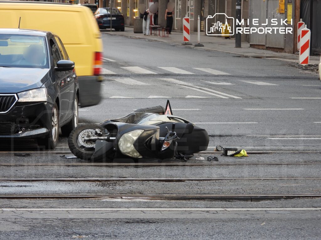 Motocykl leży na jezdni obok uszkodzonego samochodu osobowego w miejskim otoczeniu.