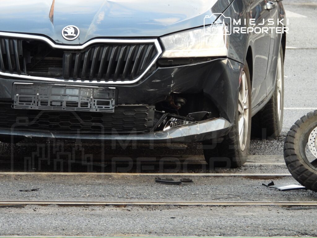 Zniszczony czarny samochód osobowy z uszkodzeniem przedniego zderzaka stoi na drodze obok torów tramwajowych.