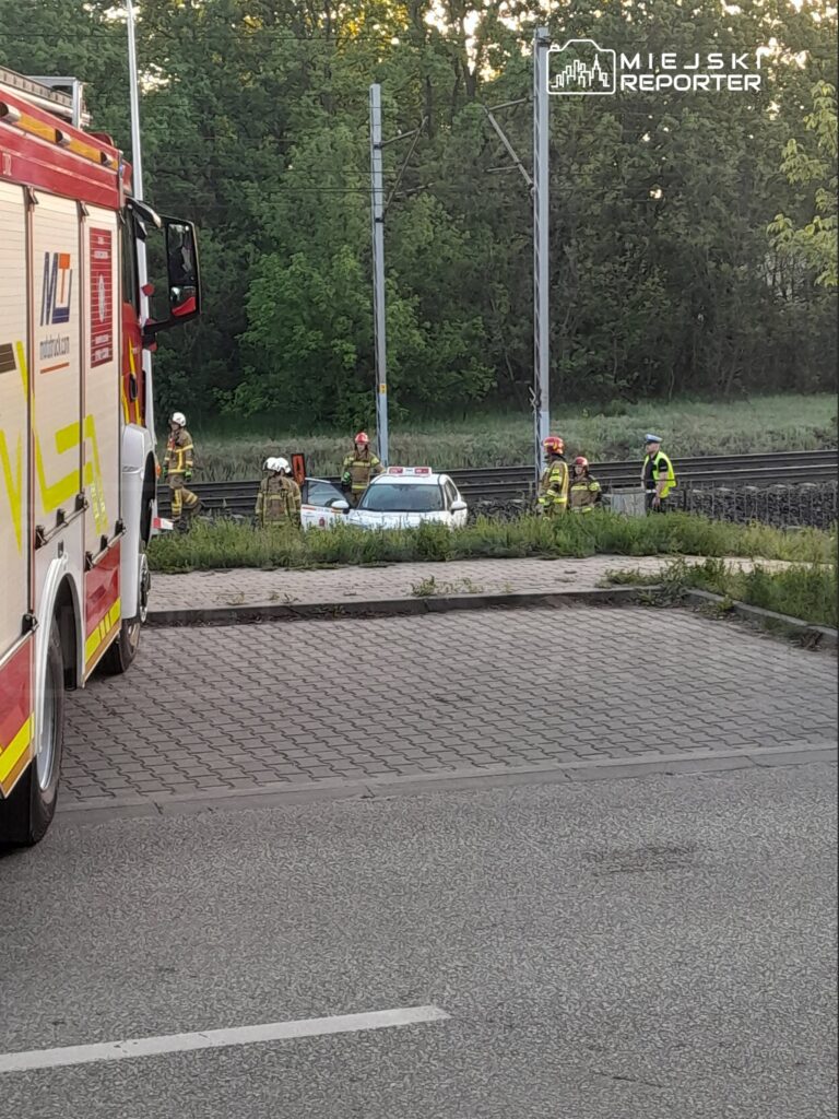 Grupa strażaków w hełmach i mundurach pracuje przy uszkodzonym samochodzie obok torów kolejowych.