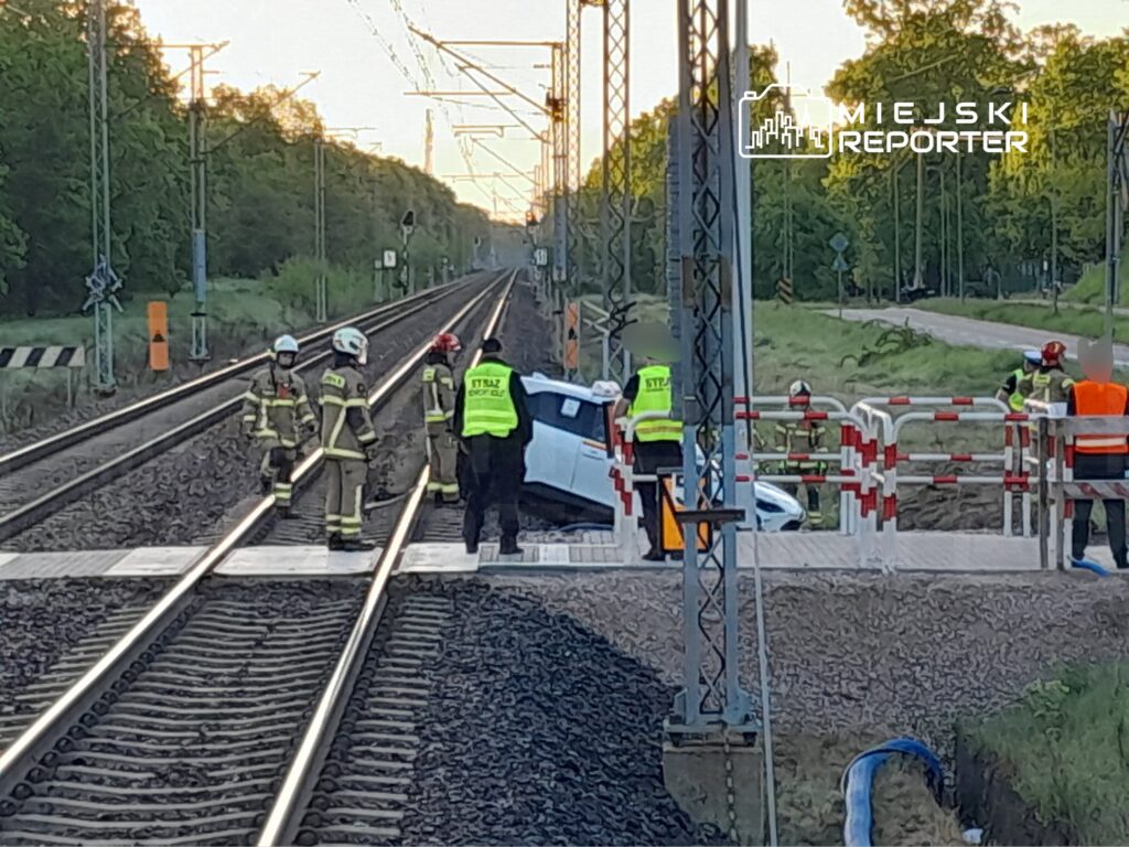 Grupa strażaków i pracowników służb drogowych pracuje przy przewróconym samochodzie na przejeździe kolejowym.