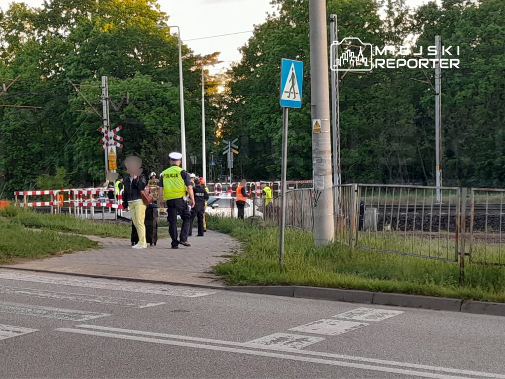 Policjanci rozmawiają z przechodniami przy przejeździe kolejowym, w tle widać zablokowaną drogę i zieleń.
