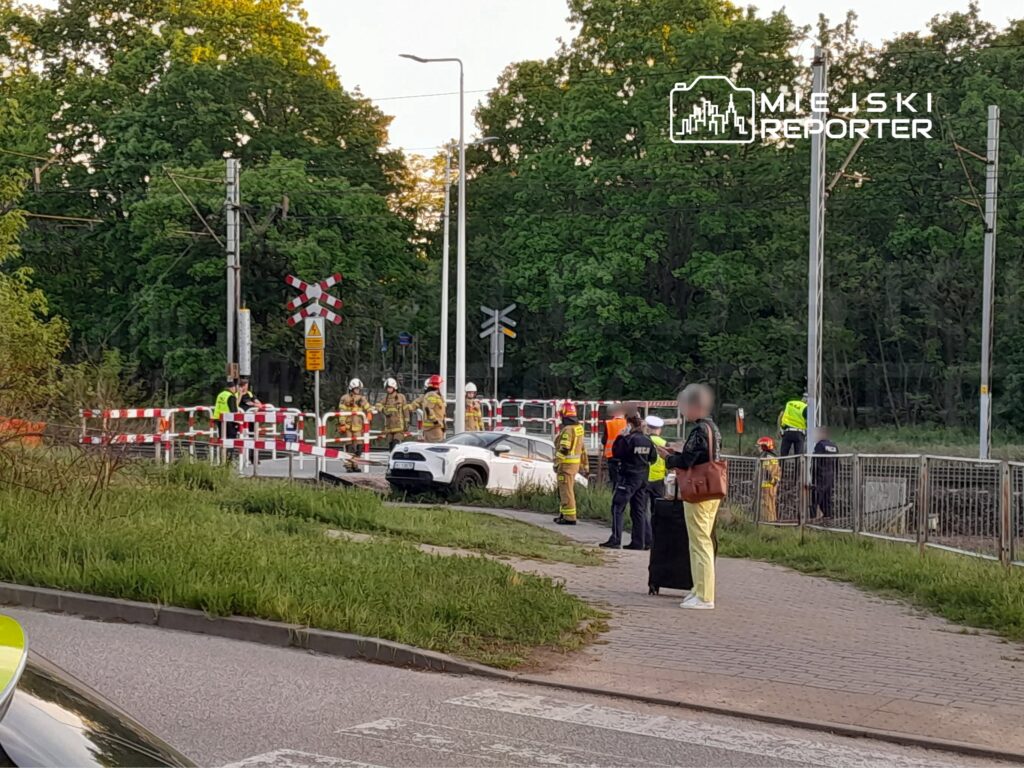 Grupa strażaków i policjantów pracuje przy zamkniętym przejeździe kolejowym, obok białego samochodu osobowego.