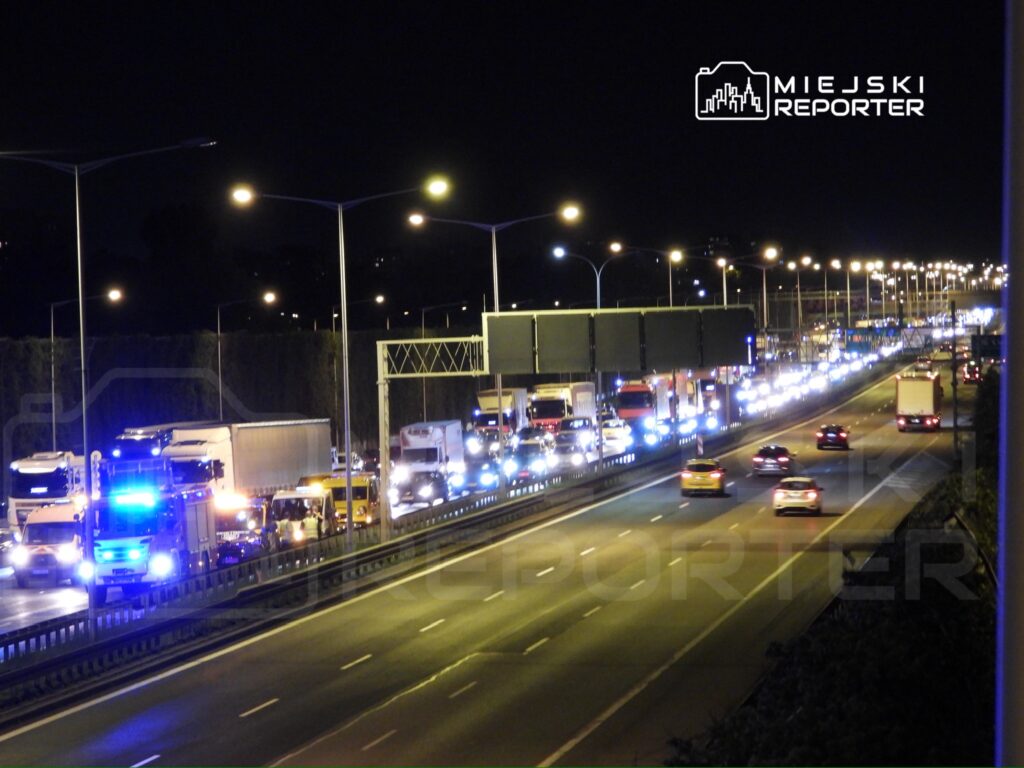 Ruch samochodowy na autostradzie w nocy, z widocznymi ciężarówkami i pojazdami osobowymi, oświetlenie uliczne w tle.