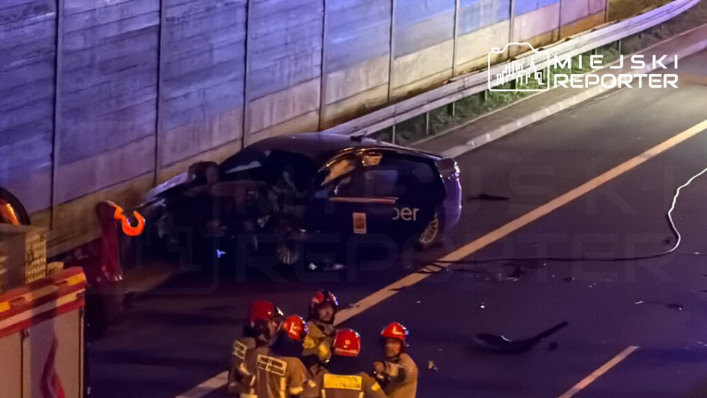 Grupa strażaków w hełmach zbiera się wokół uszkodzonego samochodu na drodze, obok widoczna jest linia policyjna.