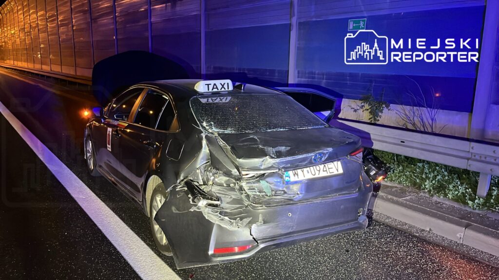 Uszkodzony samochód osobowy typu taxi z widocznymi zniszczeniami tyłu stoi na poboczu drogi, w tle nocne oświetlenie.