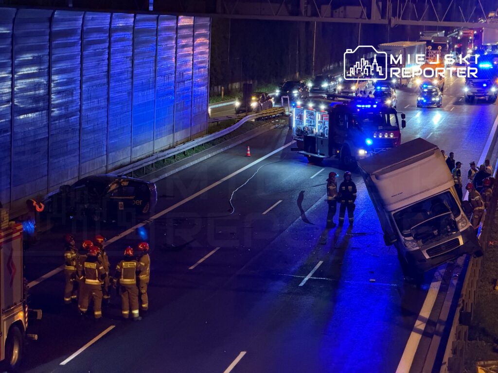 Grupa strażaków w hełmach rozmawia obok przewróconego samochodu dostawczego na autostradzie, w tle widać inne pojazdy.