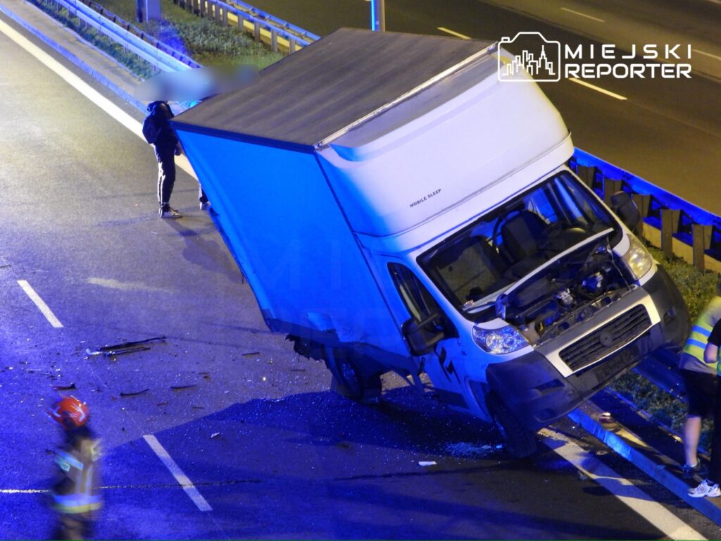 Nieoznakowany samochód dostawczy stoi na poboczu drogi, a obok niego pracują strażacy oraz mężczyzna w odblaskowej kamizelce.