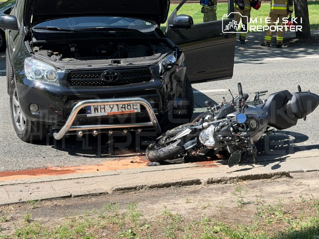Czarny samochód osobowy z otwartą maską stoi obok przewróconego motocykla, na jezdni widoczna plama płynu.