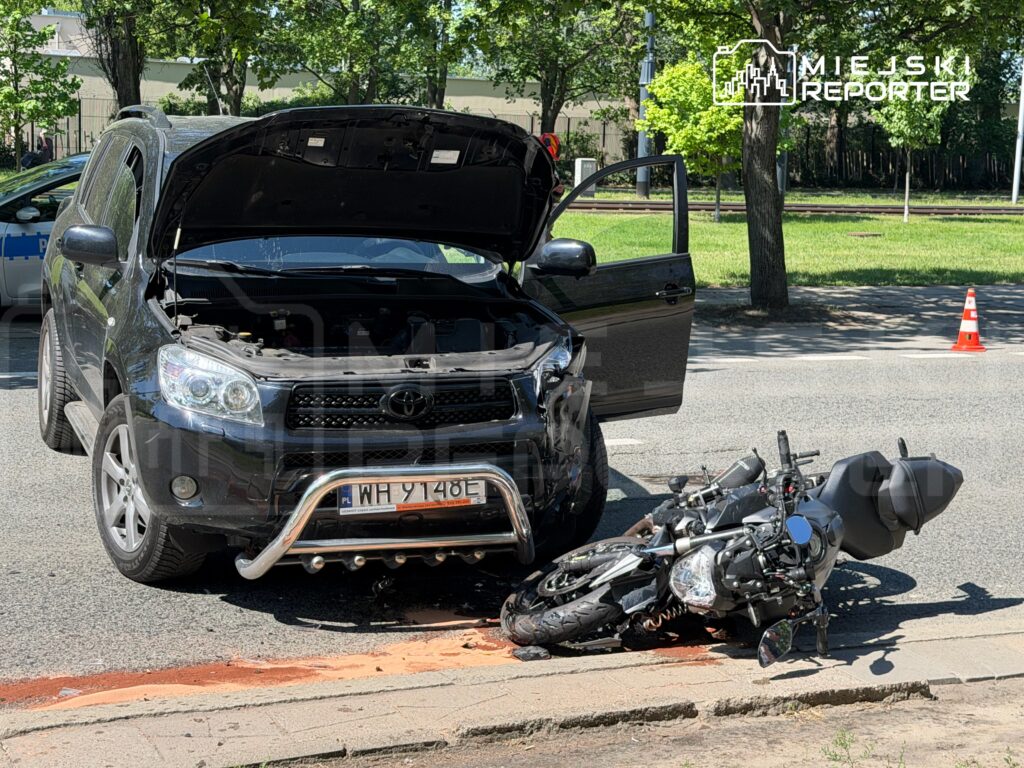 Czarny SUV z otwartą maską stoi na drodze, obok przewrócony motocykl, na jezdni widoczne plamy płynów.