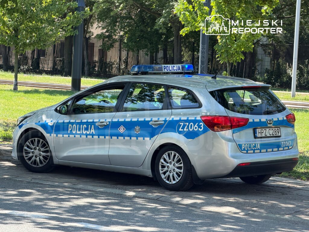 Radiowóz policyjny zaparkowany na chodniku w otoczeniu zieleni i drzew.