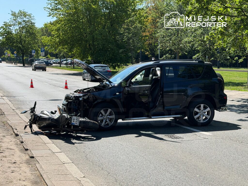 Czarny samochód osobowy z otwartą maską stoi na drodze obok przewróconego motocykla, a w tle widać zieleń drzew.