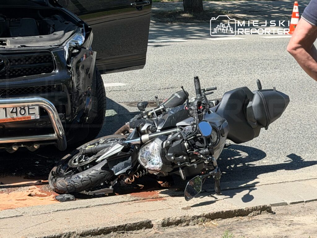 Motocykl leży na boku obok uszkodzonego samochodu osobowego na jezdni, widoczne ślady płynów.