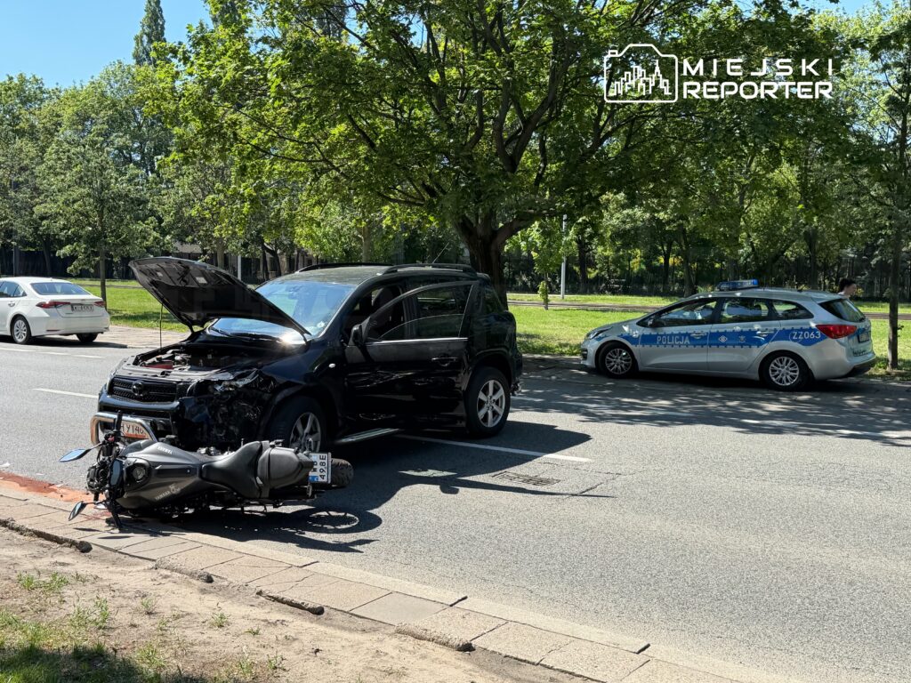 Czarny samochód osobowy z otwartą maską stoi na drodze obok przewróconego motocykla; w tle widać radiowóz policyjny.