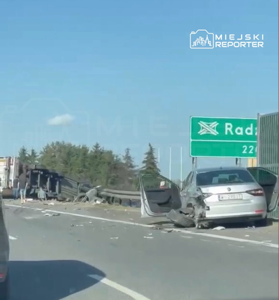 Na drodze leży przewrócony samochód ciężarowy, a obok znajduje się uszkodzone osobowe auto. W tle znak drogowy kierujący do Radzymina.