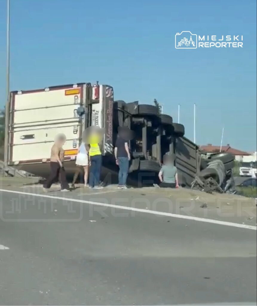 Grupa osób otacza przewrócony na bok ciężarówkę na poboczu drogi, w słoneczny dzień.