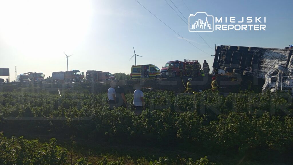 Ratownicy pracują przy przewróconym samochodzie ciężarowym na drodze, w tle widać pola i wiatraki.